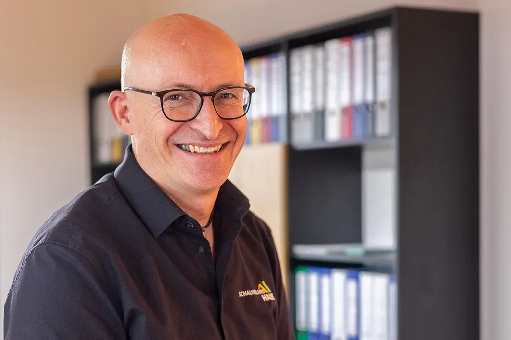 Ein kahlköpfiger Mann mit Brille lächelt in einem schwarzen Hemd vor einem Bücherregal voller Ordner.