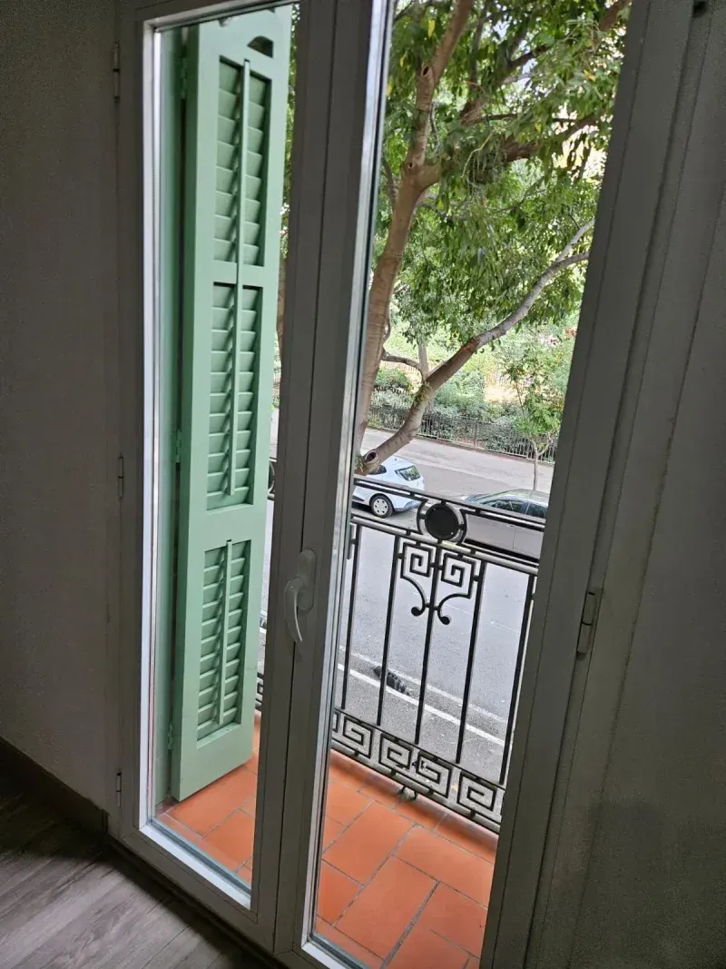 Puerta abierta a balcón con persianas verdes, barandilla de hierro y vista a la calle.