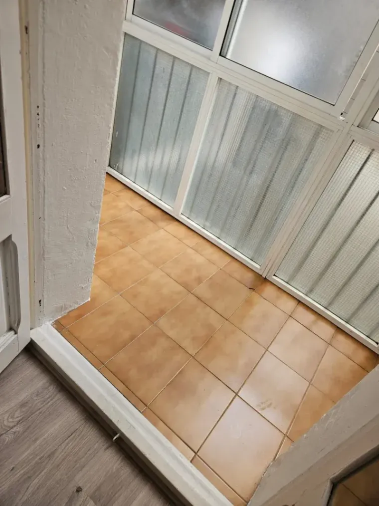 Vista de un pequeño porche cerrado y embaldosado con marco de ventana y puerta de color blanco.