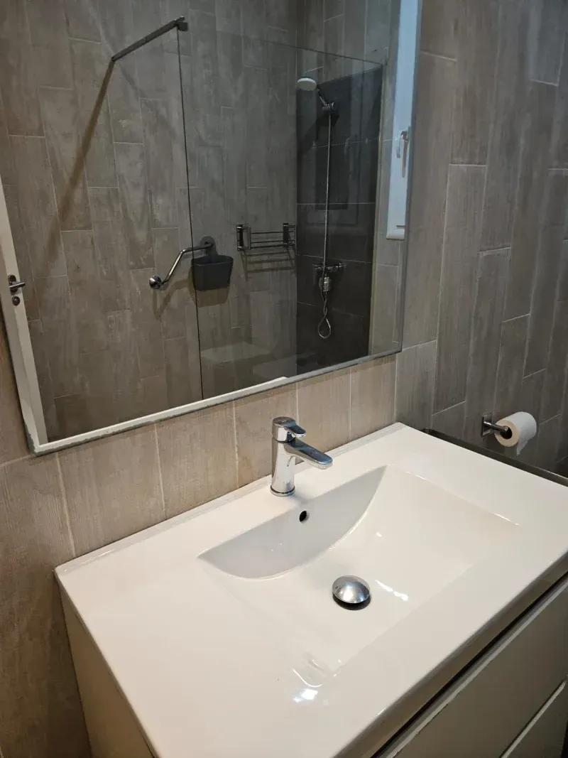 Baño con lavabo blanco, espejo grande y ducha reflejada. Paredes de azulejos grises.