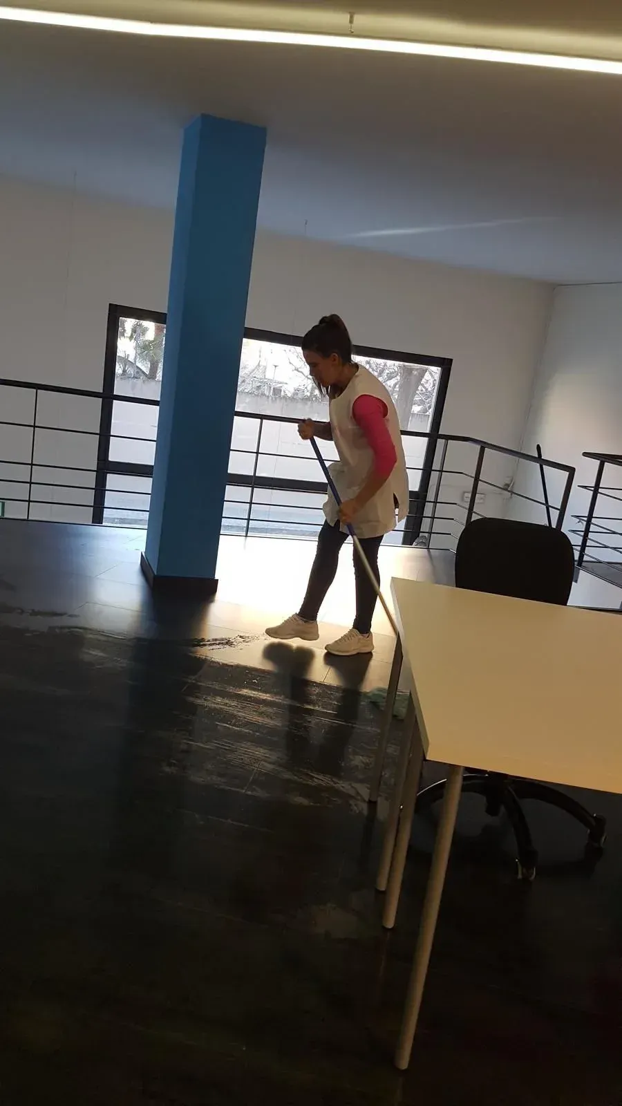 Persona barriendo un piso oscuro, gran pilar azul, paredes blancas y escritorio.