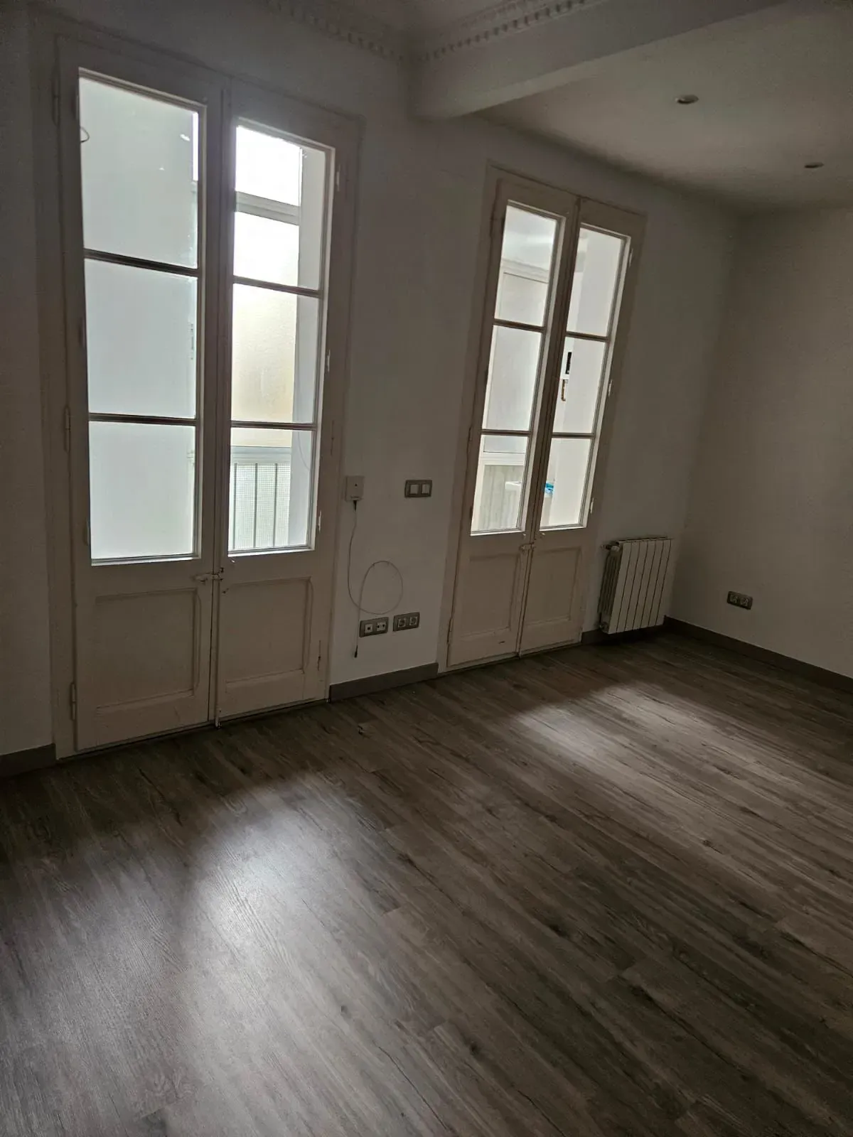 Habitación vacía con suelo de madera, dos puertas altas blancas y un pequeño radiador.