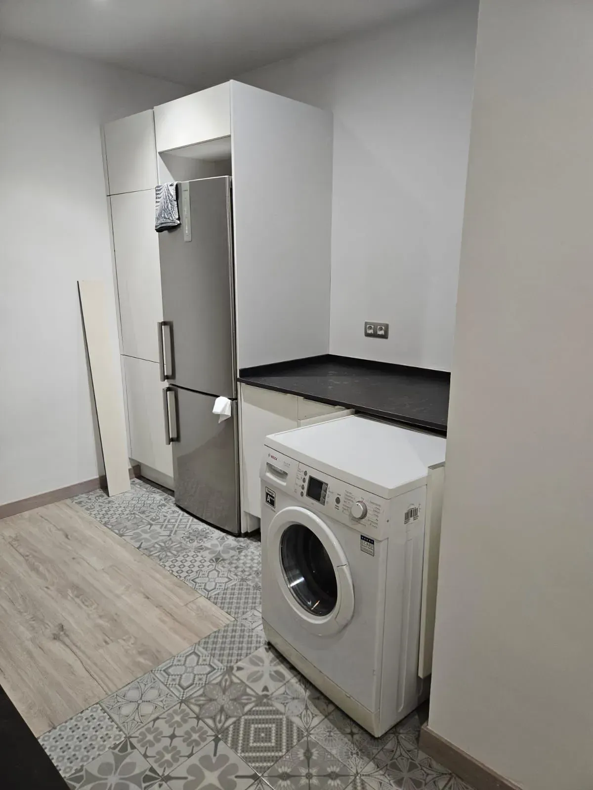 Una pequeña cocina con refrigerador, lavadora y armarios. Paredes claras y suelo estampado.