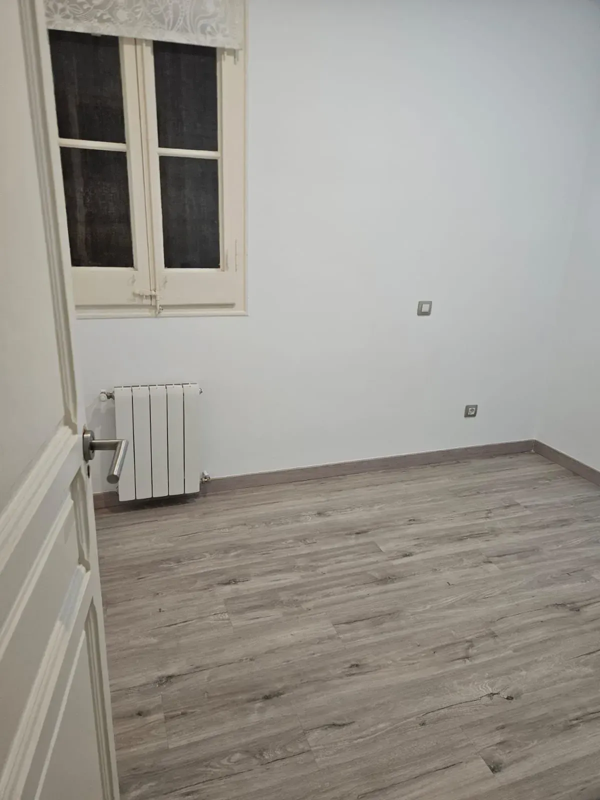 Habitación vacía con paredes blancas, suelo imitación madera clara, ventana, radiador y una puerta parcialmente abierta.