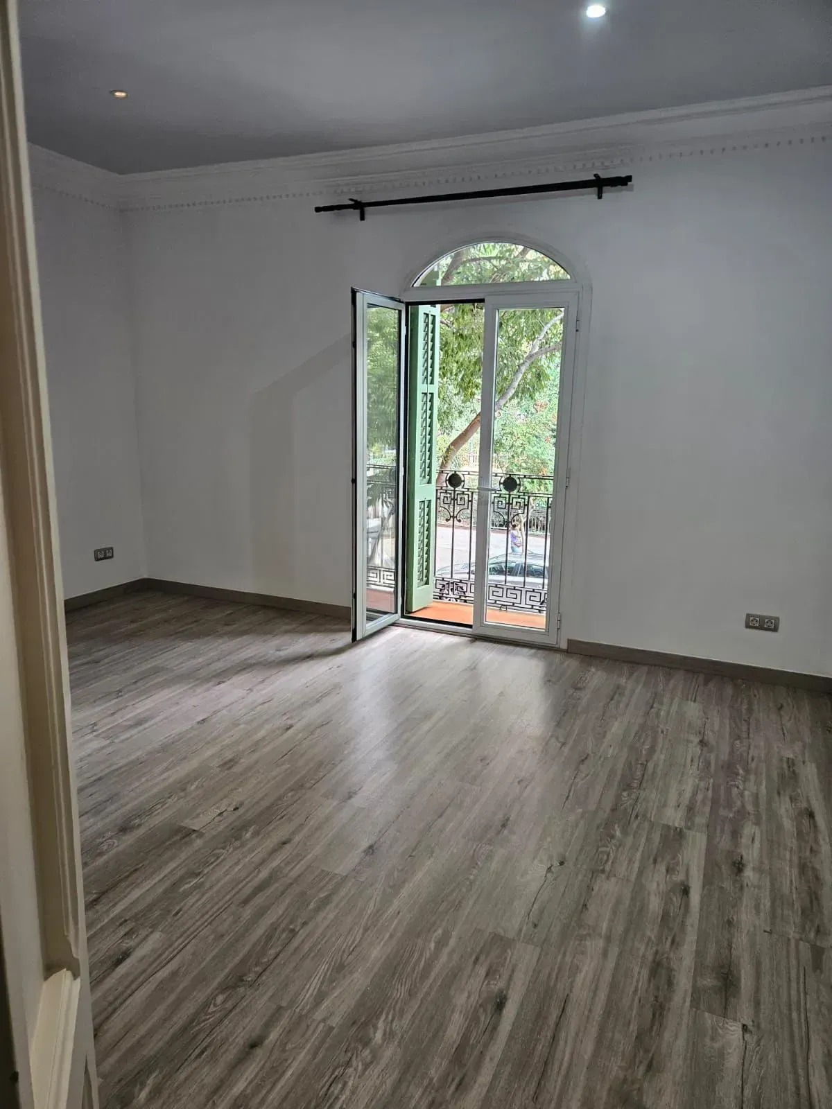Habitación vacía con pisos de madera, paredes blancas y una puerta de balcón con vista a la vegetación.