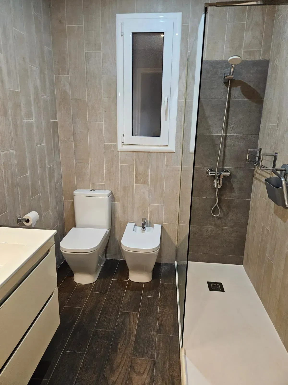 Baño con inodoro, bidé, ducha y tocador. Paredes de azulejos beige, suelo de imitación madera oscura, ventana