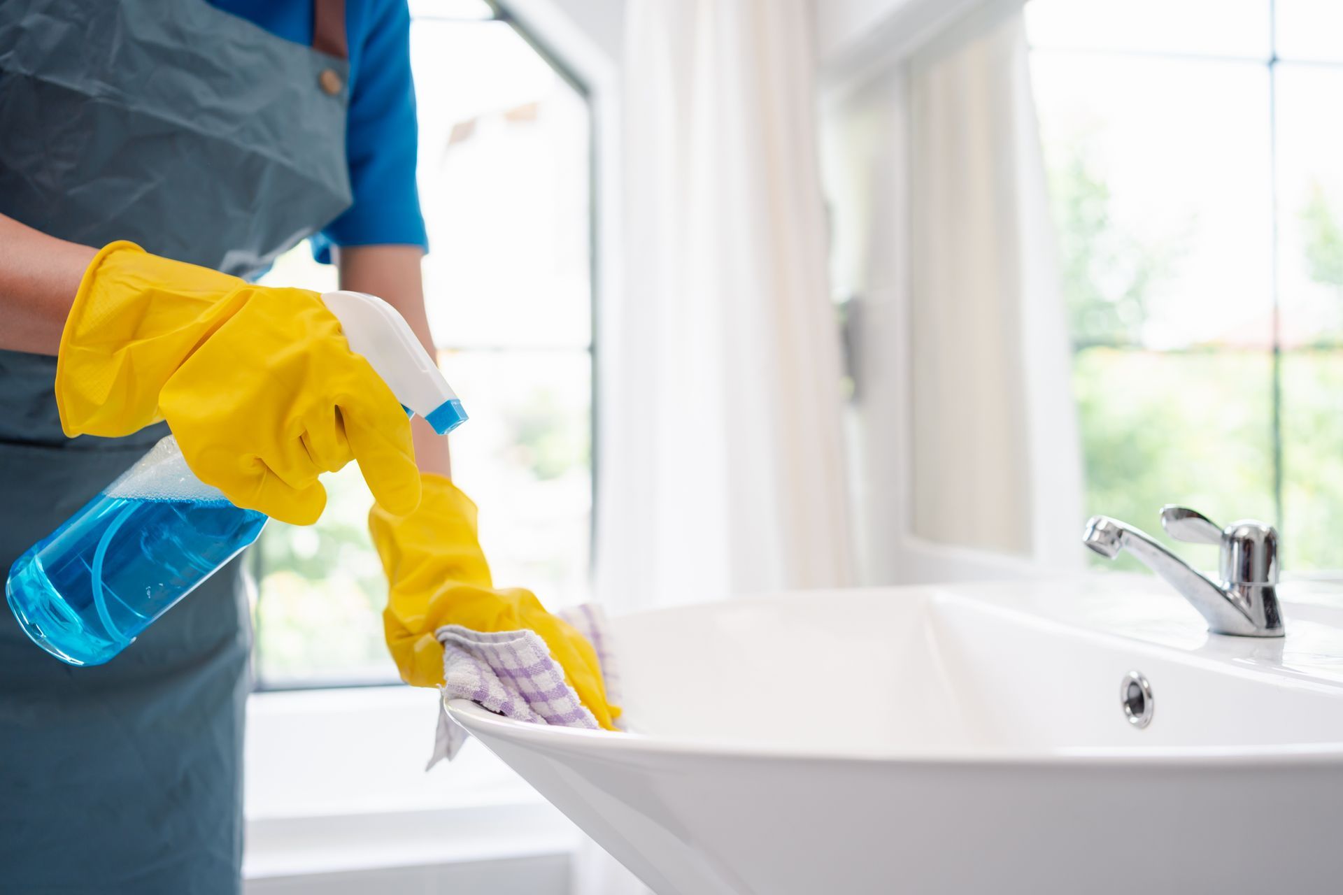 Une personne portant des gants jaunes nettoie un lavabo avec un nettoyant bleu en spray et un chiffon.