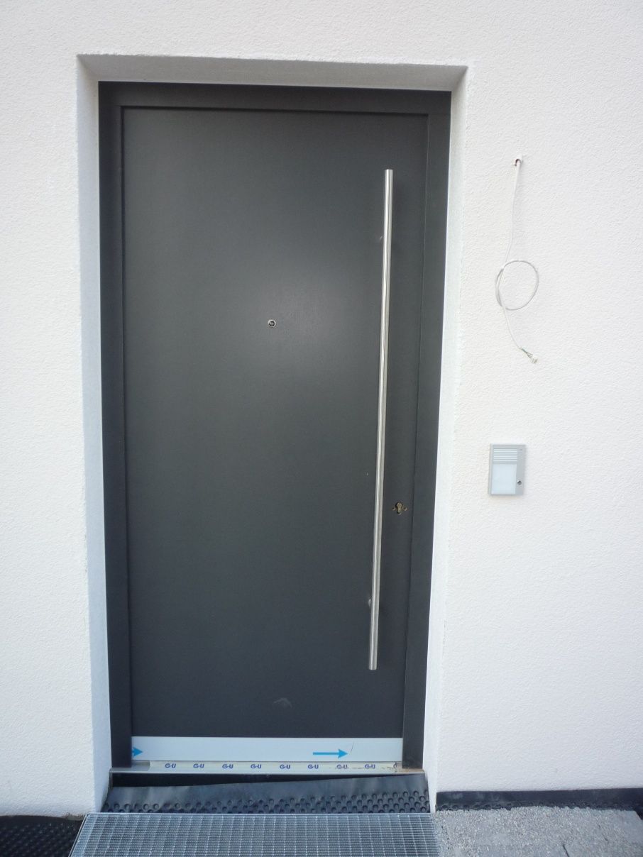 Dunkelgraue Eingangstür mit langem, vertikalem Griff und Guckloch, in eine weiße Wand eingelassen.