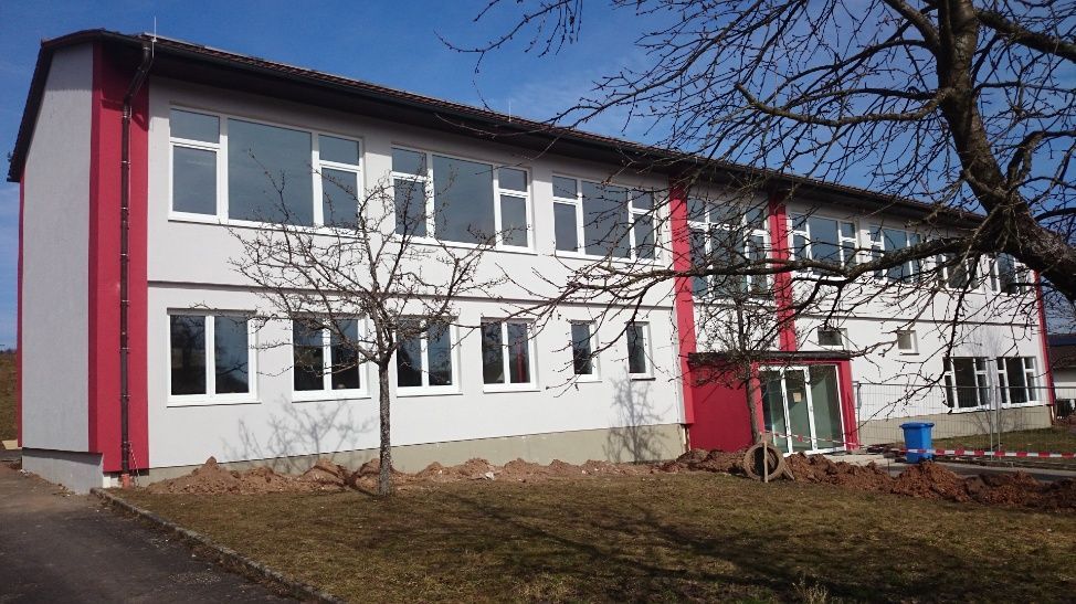 Zweistöckiges Gebäude mit weißen Wänden, roten Akzenten und vielen Fenstern, möglicherweise eine Schule.