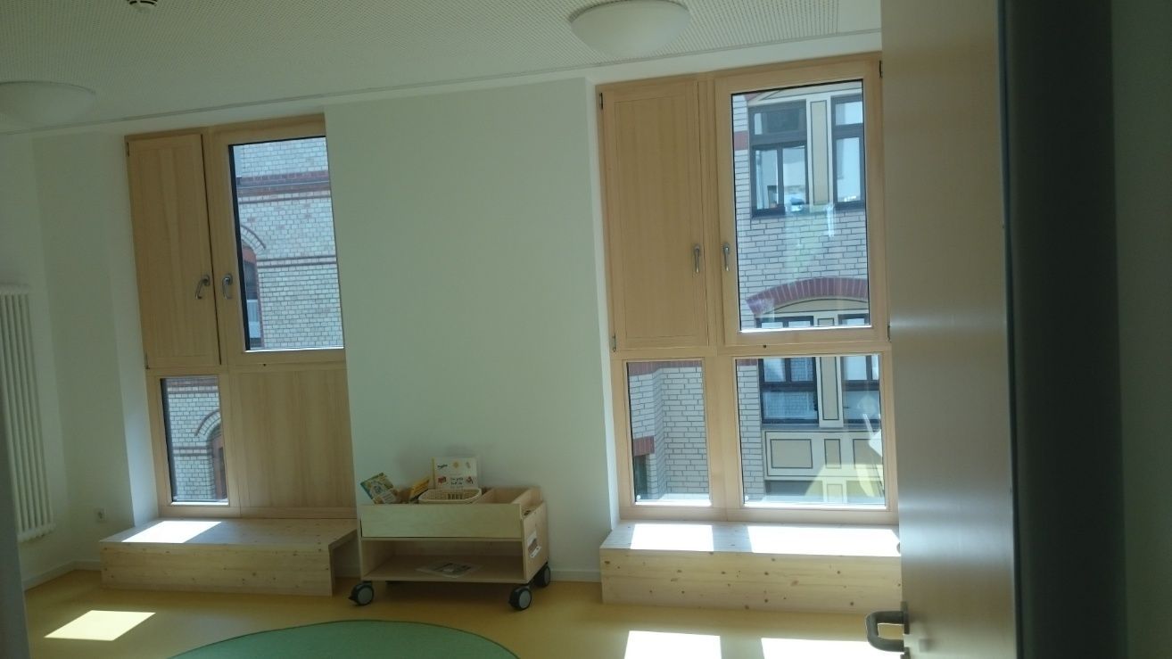 Heller Raum mit zwei Fenstern, hellen Holzrahmen und Fensterläden, einem Rollwagen und einer runden Matte auf dem Boden.