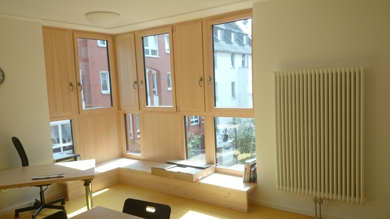Gemütliches Eckzimmer mit großen Fenstern und Blick auf die Gebäude. Mit Heizkörper, Schreibtisch und eingebauter Bank.