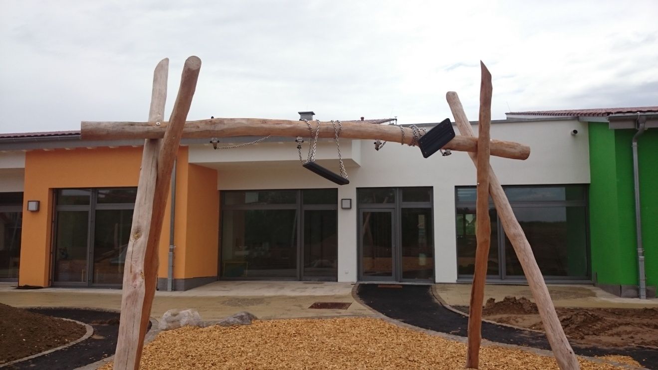 Holzschaukel vor einem farbenfrohen Gebäude mit Fenstern, auf einem Spielplatz.