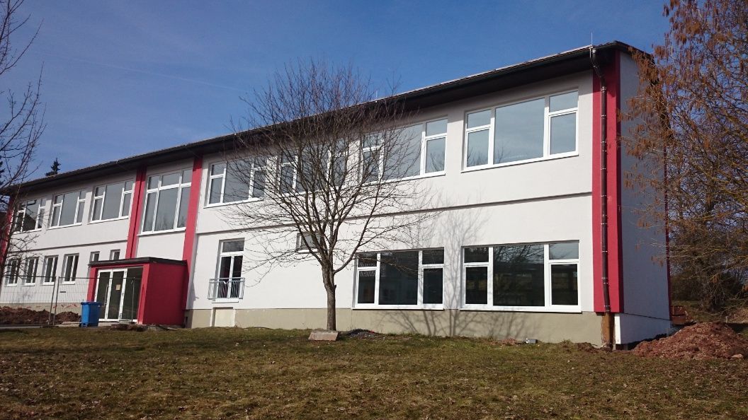 Zweistöckiges weißes Gebäude mit roten Akzenten und großen Fenstern; kahler Baum davor, blauer Himmel.