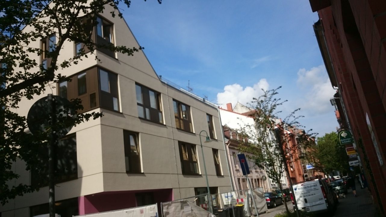 Modernes cremefarbenes Gebäude mit mehreren Fenstern vor blauem Himmel, neben einem roten Backsteingebäude in einer Straße.