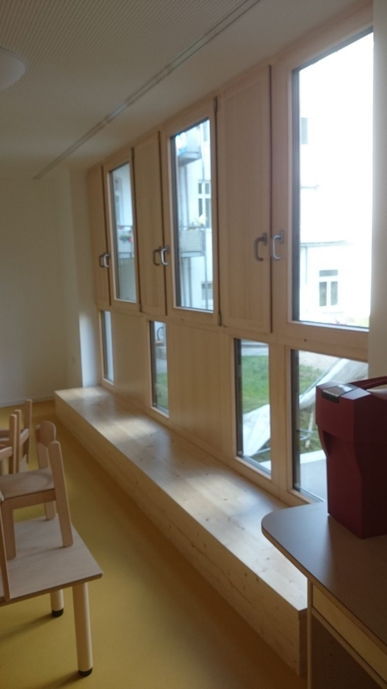 Eine Reihe großer Fenster in einem Raum, darunter eine eingebaute Holzbank und ein kleiner Tisch.