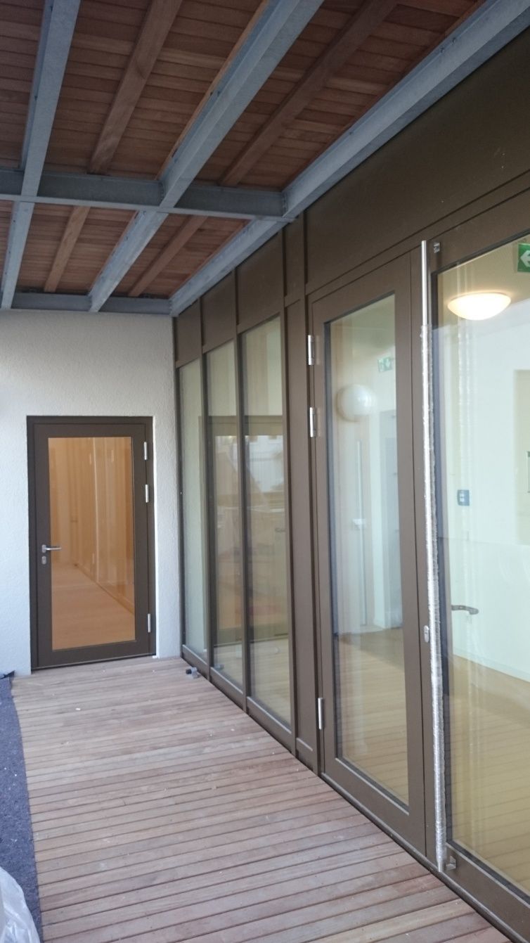 Holzsteg führt zu Glastüren und einer braunen Tür. Die Decke ist mit Holzpaneelen versehen.