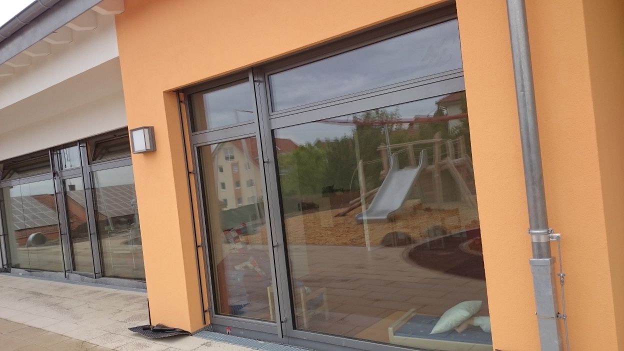 Außenansicht des Gebäudes mit orangefarbenen Wänden und großen Fenstern, in denen sich ein Spielplatz widerspiegelt.
