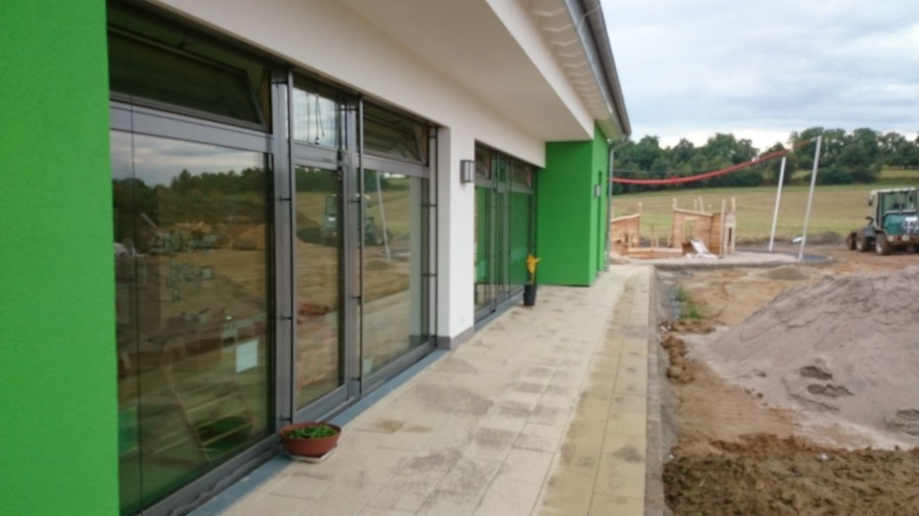 Grünes Gebäude mit großen Fenstern, einem Betonweg und einer Baustelle im Hintergrund.