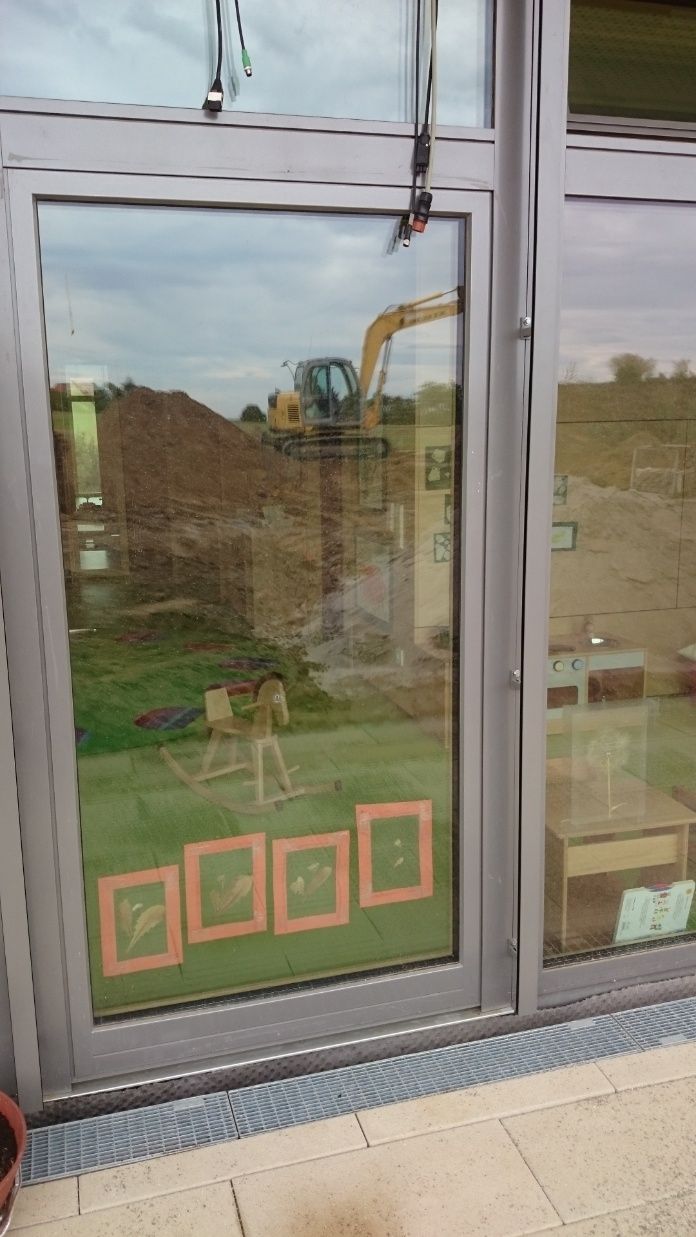 Bagger spiegelt sich in einer Glastür; Baustelle mit Rasenfläche und orangefarbenen Rahmen.