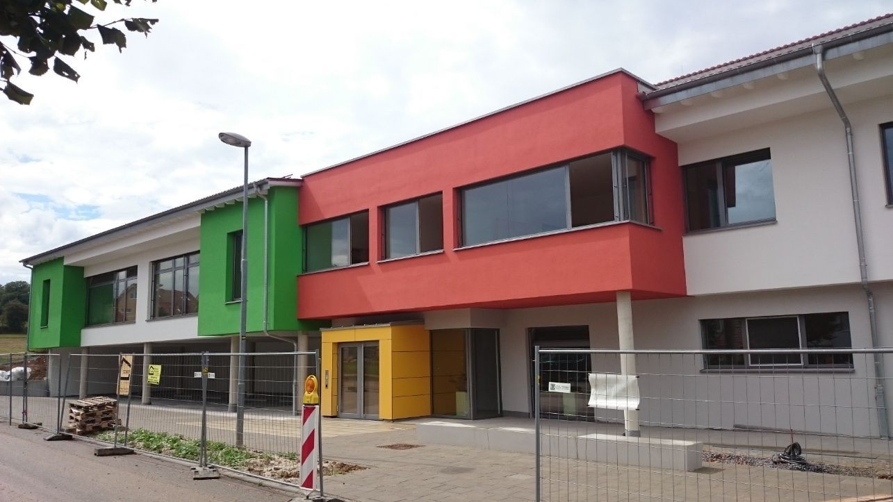 Ein modernes Gebäude mit roten, grünen und weißen Abschnitten, im Bau mit einem gelben Eingang, hinter einem Bauzaun.