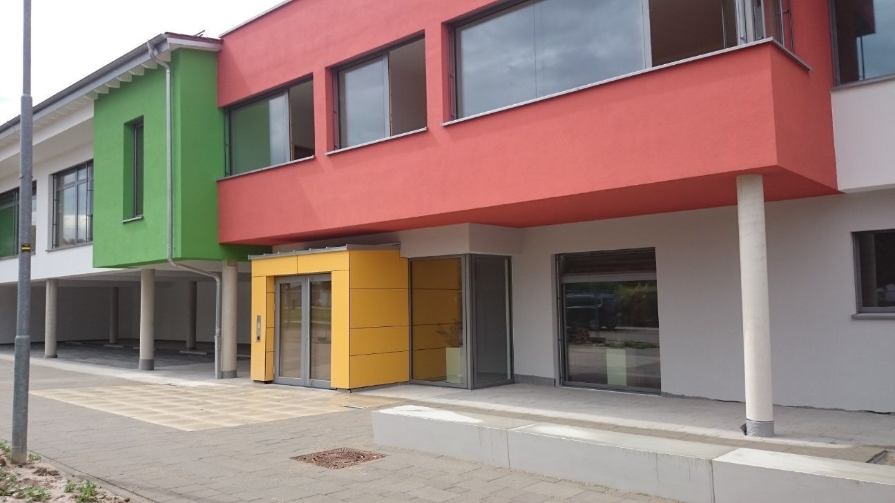 Gebäude mit farbenfroher Fassade; grüne, rote und gelbe Bereiche. Eingangsbereich und Säulen tragen ein zweites Stockwerk.