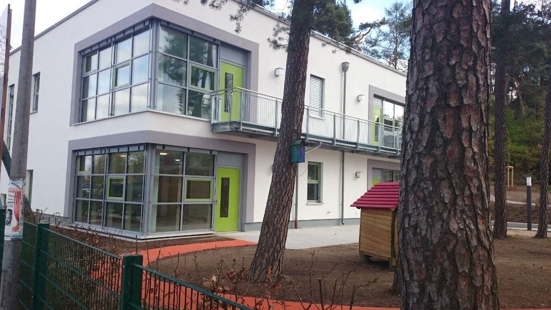 Modernes zweistöckiges Gebäude mit großen Fenstern und lindgrünen Türen, umgeben von Bäumen und einem roten Weg.