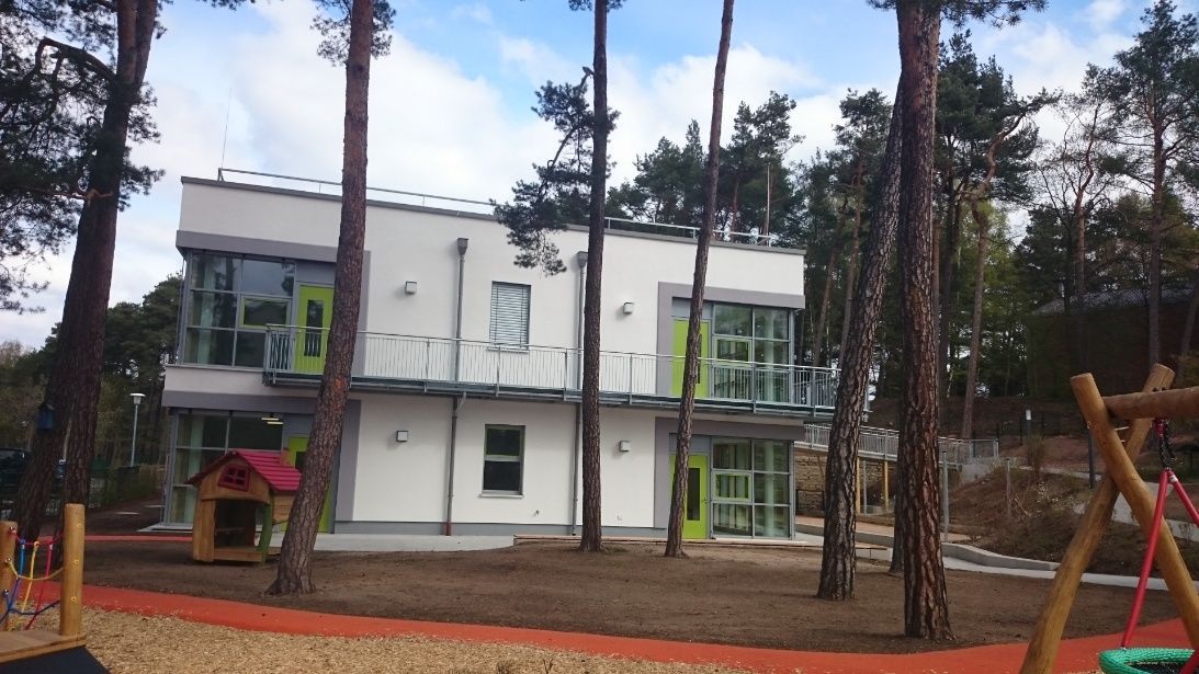 Weißes zweistöckiges Gebäude mit großen Fenstern zwischen Bäumen. Spielplatz davor mit rotem Gehweg.