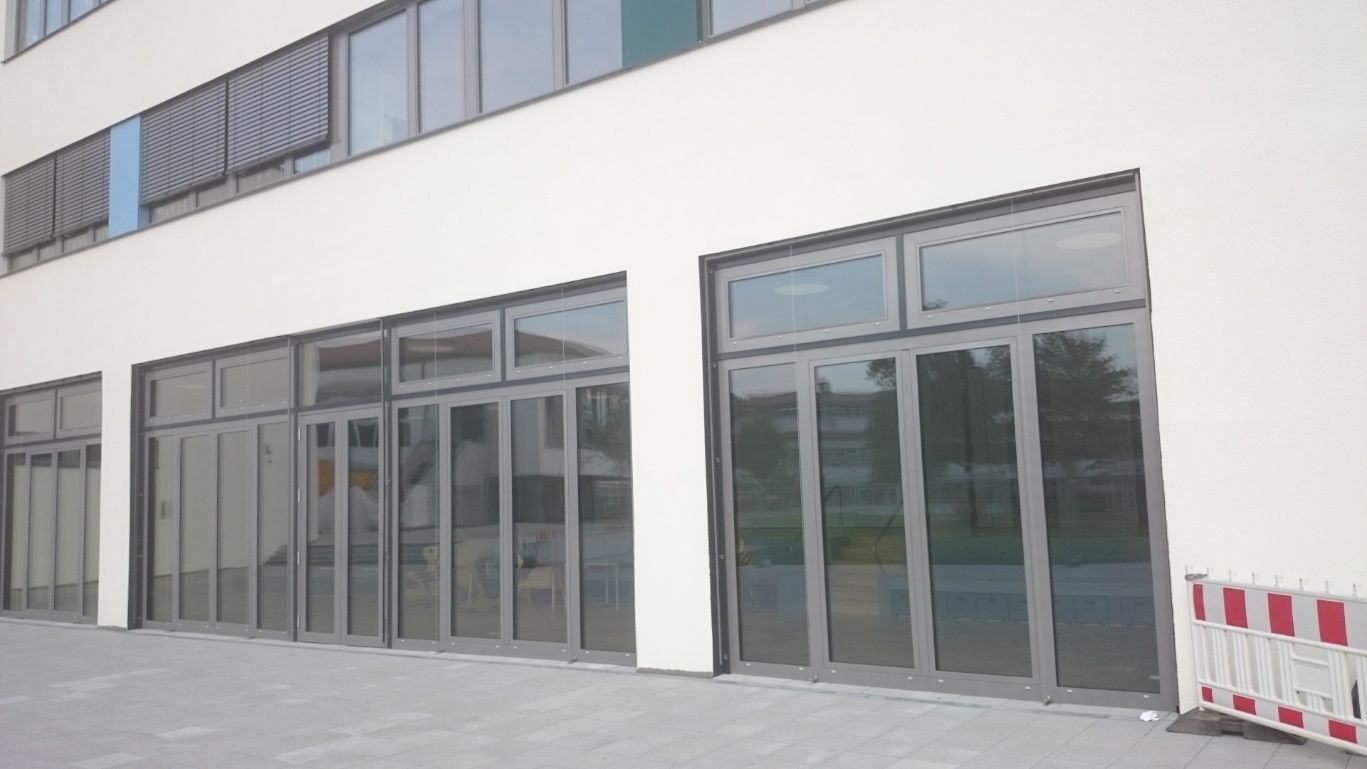 Außenansicht eines Gebäudes mit großen Glastüren und -fenstern, grauen Rahmen, weißen Wänden und einem grauen Gehweg.