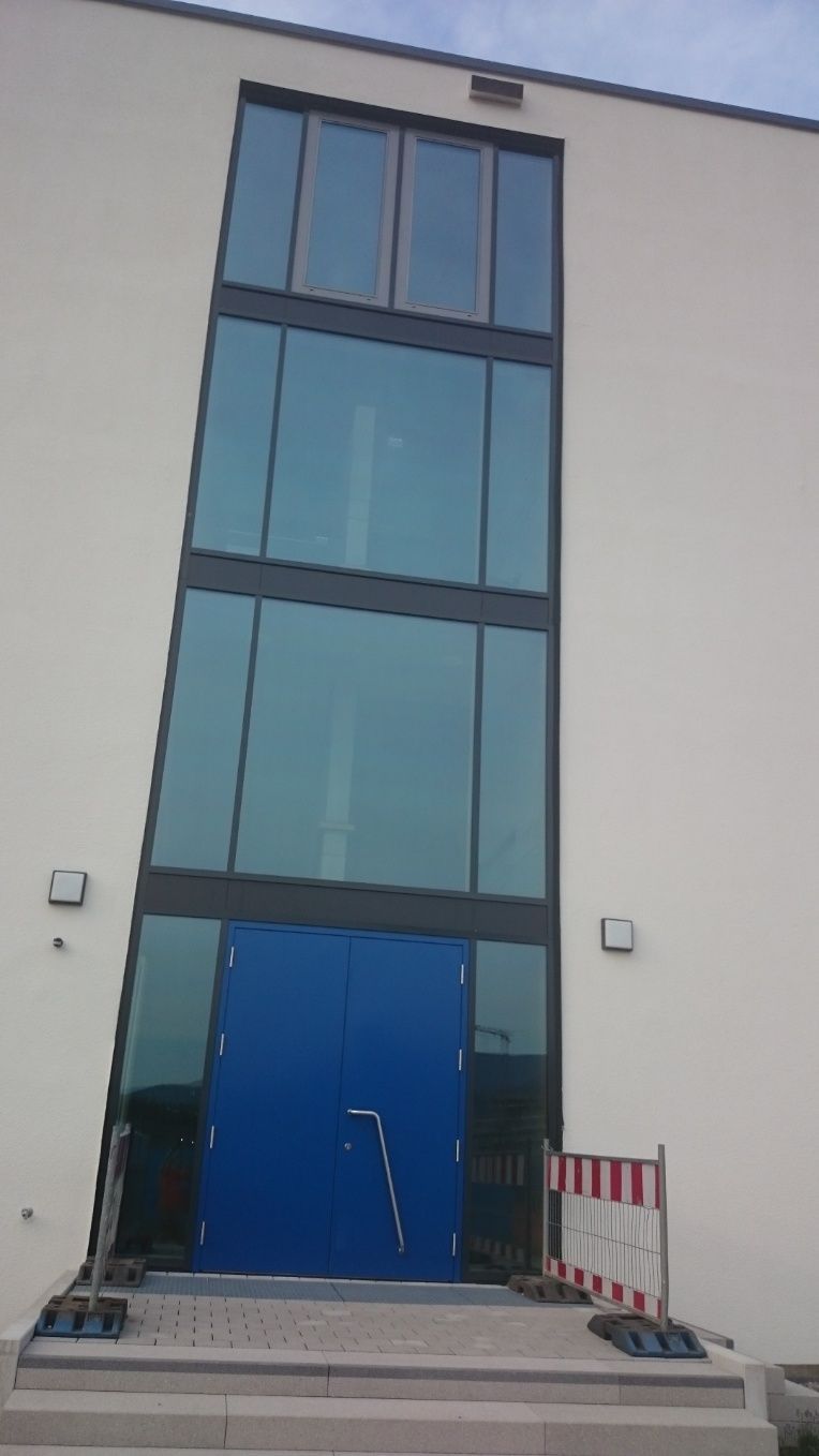 Außenansicht eines Gebäudes mit blauen Türen und großen Glasfenstern. Auf der rechten Seite befindet sich eine rot-weiße Schranke.