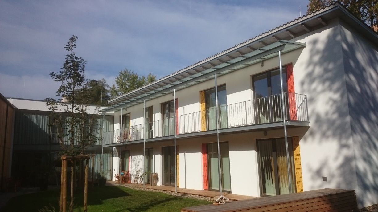 Weißes zweistöckiges Gebäude mit Balkonen, bunten Türrahmen und einem kleinen grünen Rasen.