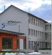 Schulgebäude mit weißen und orangefarbenen Wänden; Logo „Suntalachschule“.