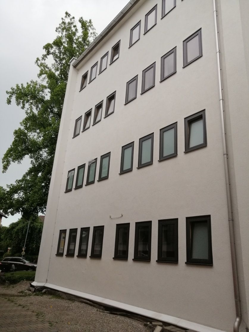 Weißes Gebäude mit Reihen dunkel gerahmter Fenster unter einem bewölkten Himmel, links Bäume.