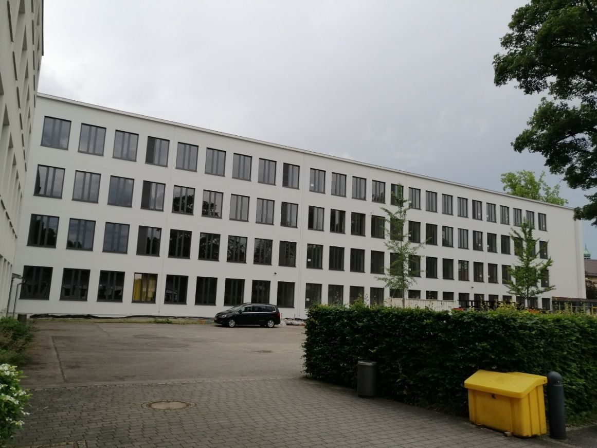 Weißes mehrstöckiges Gebäude mit vielen rechteckigen Fenstern. Schwarzes Auto davor geparkt. Bewölkter Tag.