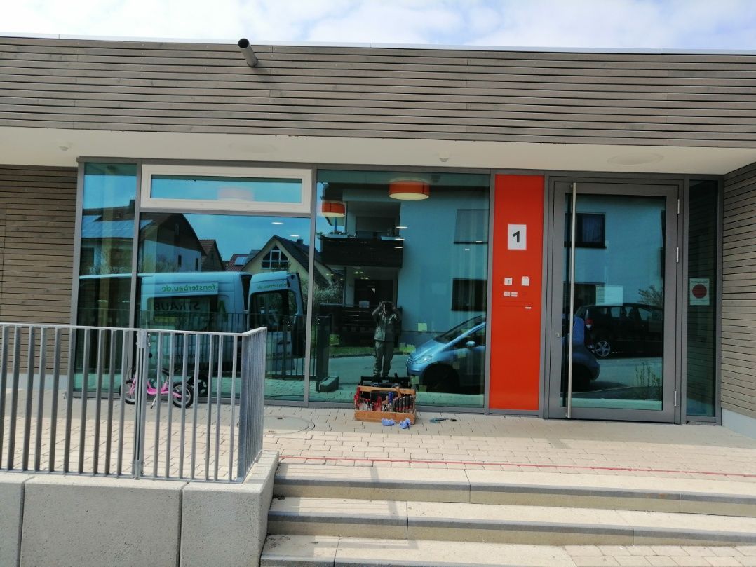 Außenansicht eines Gebäudes mit Glasfenstern, einer roten Tür und einer Betonrampe. Spiegelungen zeigen eine Straßenszene.