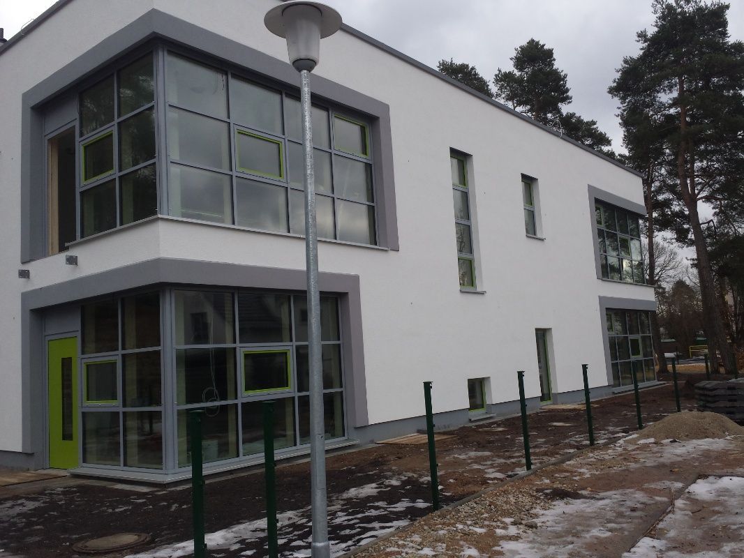 Modernes weißes Gebäude mit großen Fenstern und grauen Zierleisten, einer grünen Tür und einem Wald im Hintergrund.