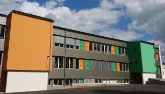 Schulgebäude mit grauer Fassade, bunten orangefarbenen und grünen Akzenten vor einem teilweise bewölkten Himmel.