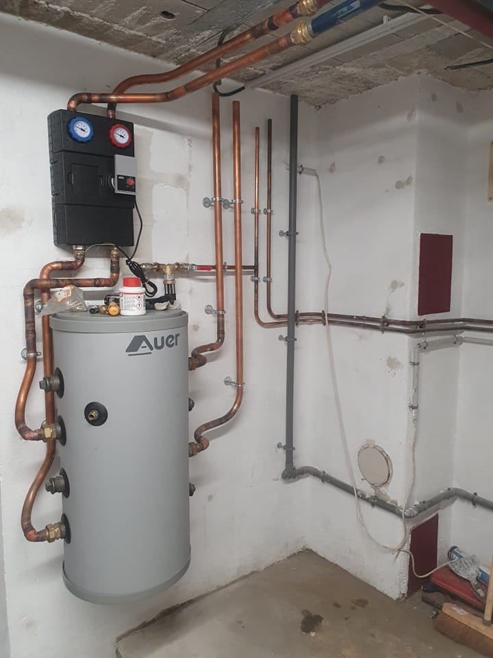 Chauffe-eau gris et unité de commande noire avec tuyaux en cuivre sur fond de murs blancs.