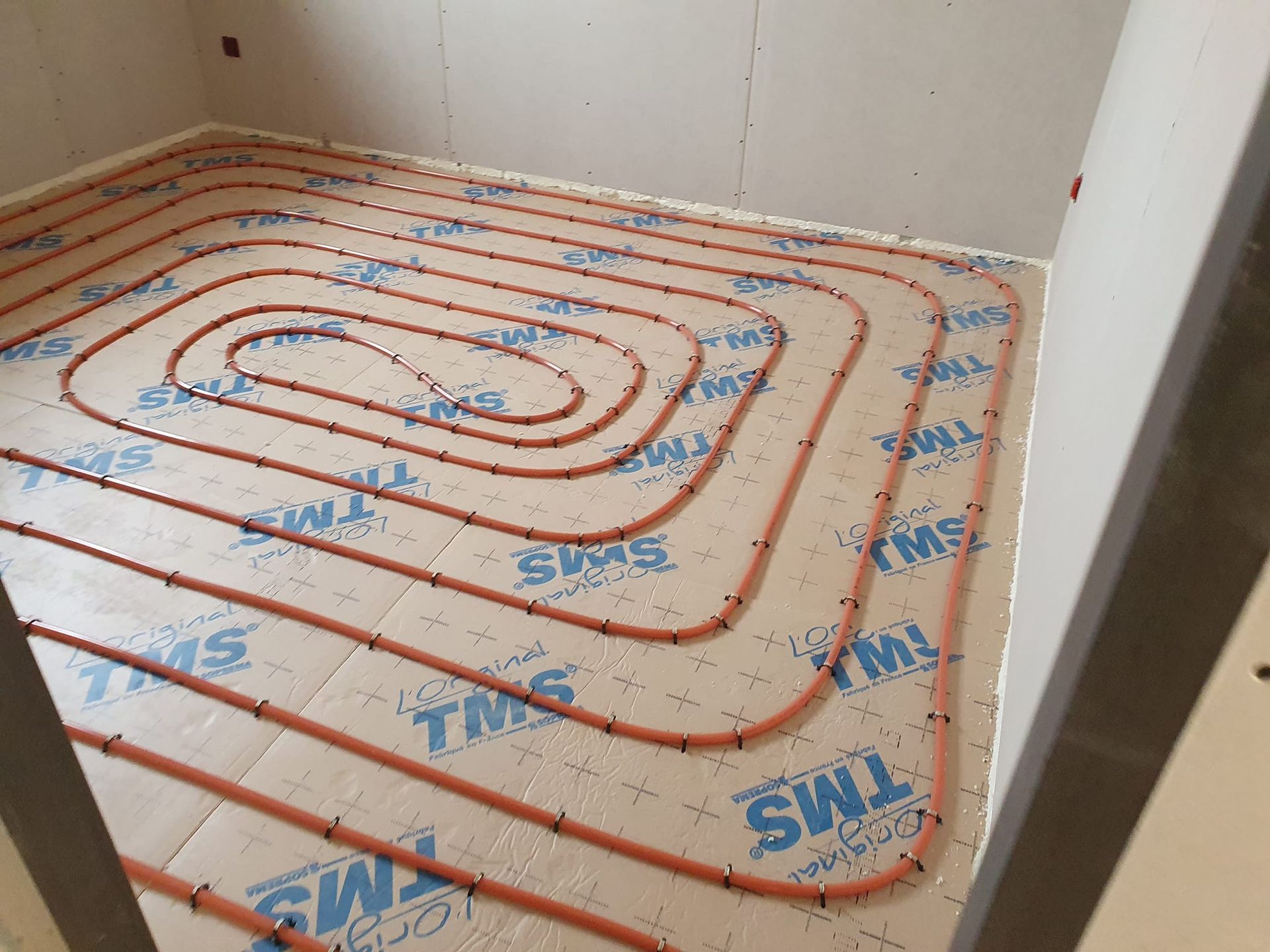 Des tuyaux orange disposés en spirale sur le sol, au-dessus d'un isolant bleu et blanc ; une pièce en construction.