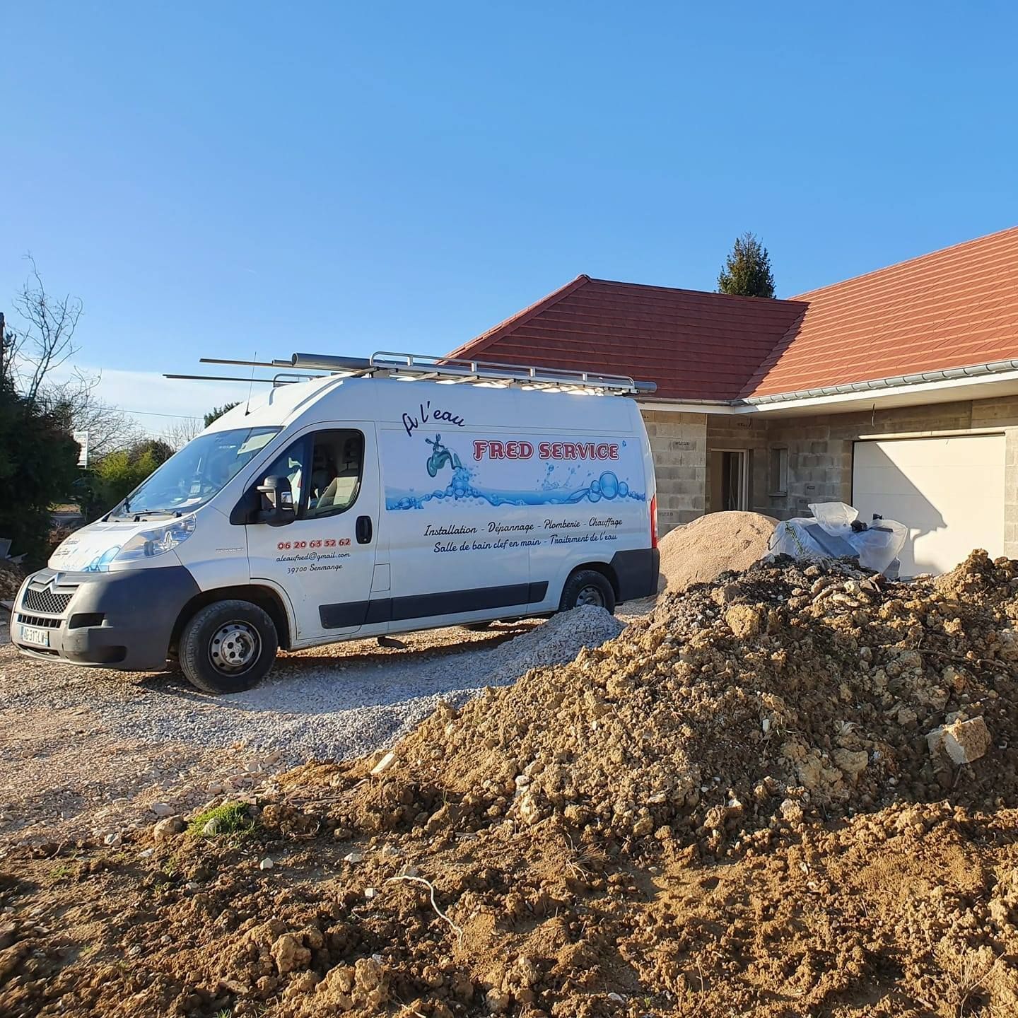Camionnette de chantier blanche avec logo, garée devant une maison en construction ; tas de terre au premier plan.