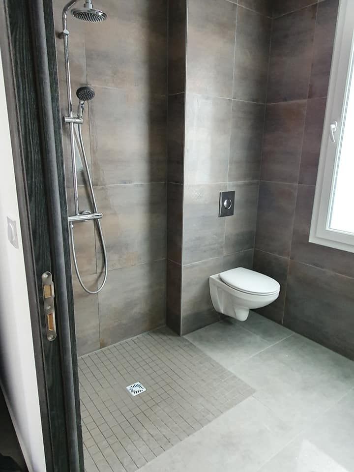 Cabine de douche avec carrelage gris, toilettes suspendues et fenêtre dans une salle de bains moderne.
