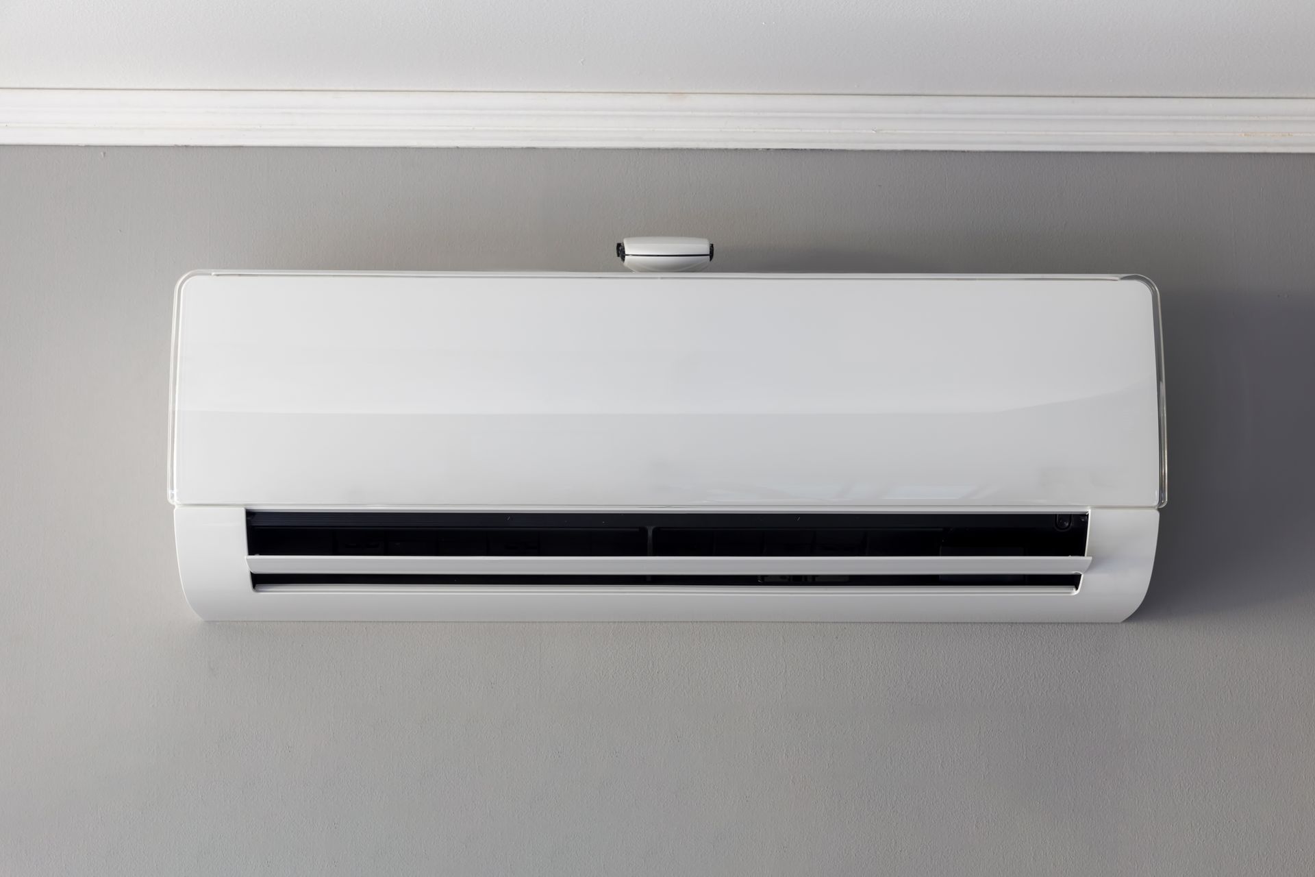 Climatiseur blanc sans conduit, fixé sur un mur gris, près d'une moulure de plafond blanche.
