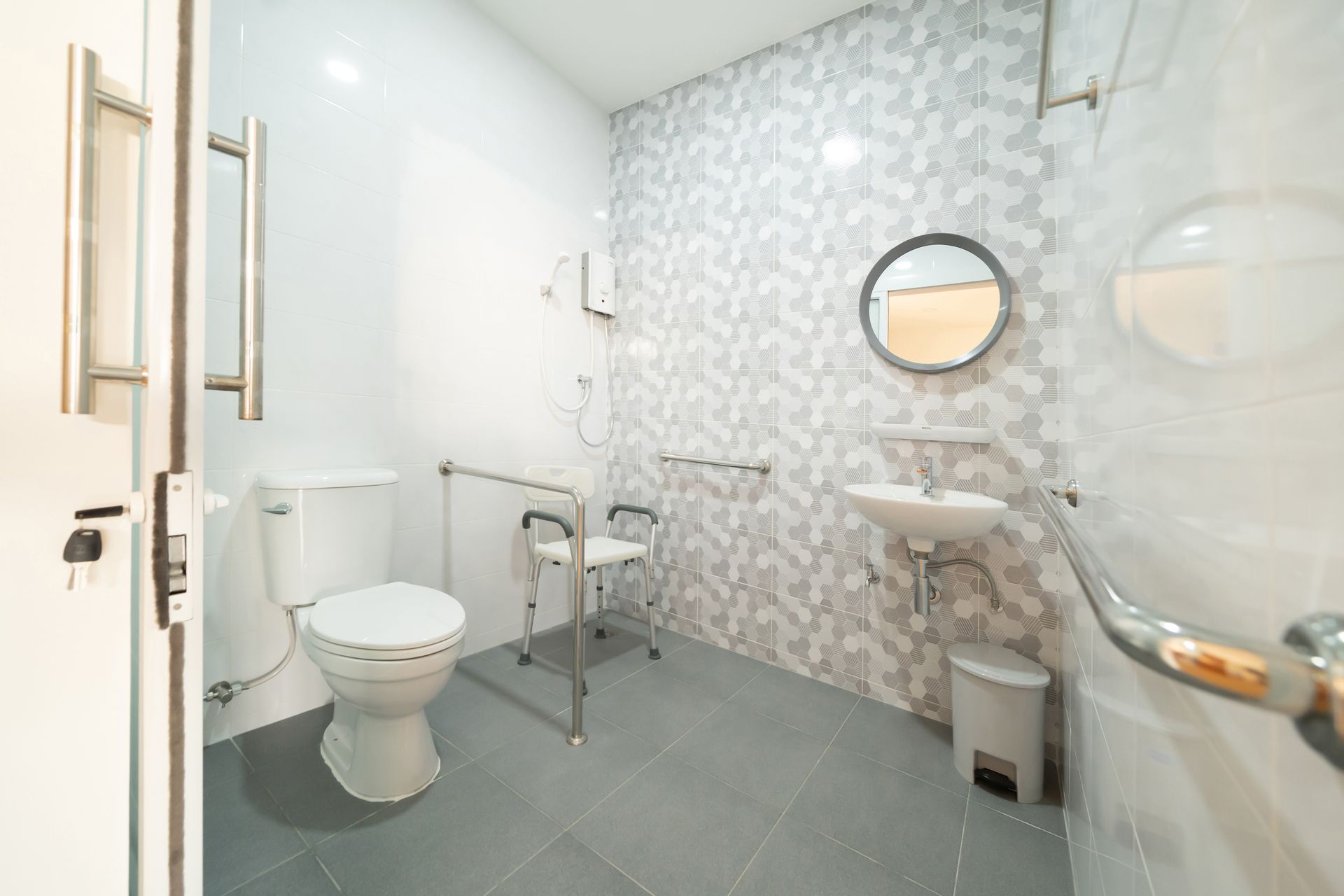 Salle de bains accessible avec toilettes, lavabo, barres d'appui et douche, carrelage gris et blanc, siège de douche.