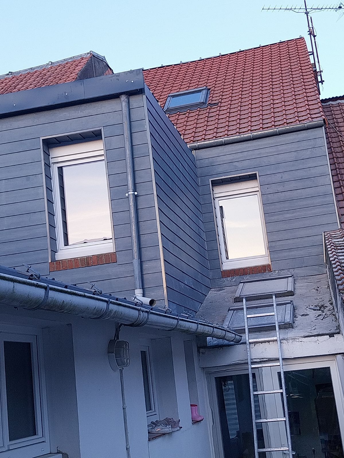 Bâtiment aux façades grises avec deux fenêtres, un toit en tuiles rouges et une échelle.