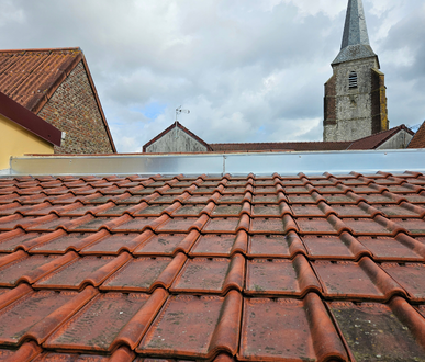 Toit de tuiles rouges, avec un clocher d'église et un ciel nuageux en arrière-plan.