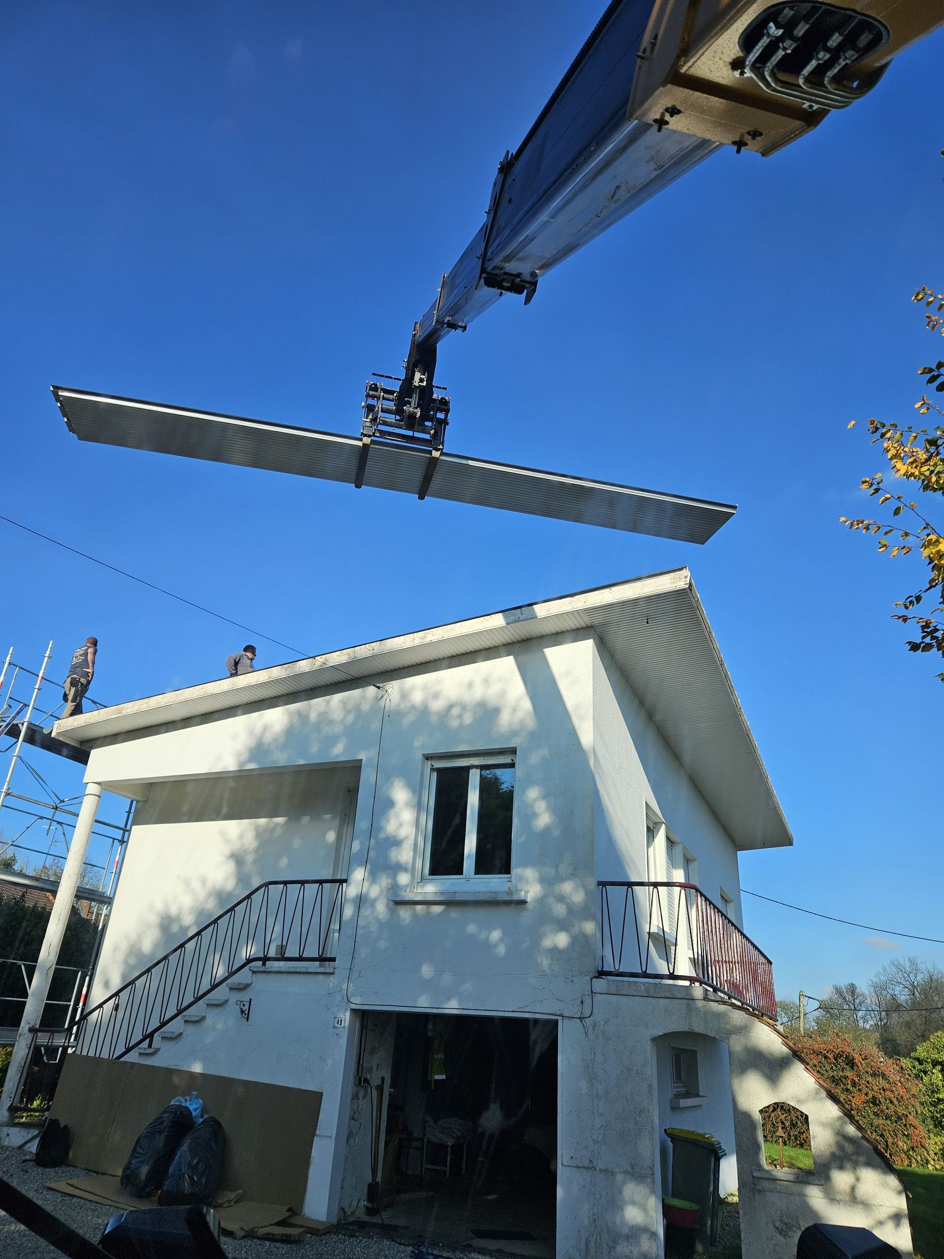 Une grue soulève une grande structure métallique au-dessus d'une maison blanche ; des ouvriers sont sur le toit.
