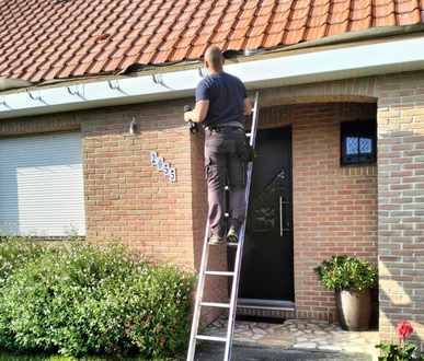 Un homme sur une échelle installe quelque chose sur l'extérieur de la maison, façade en briques, porte sombre, verdure et toit en tuiles rouges.