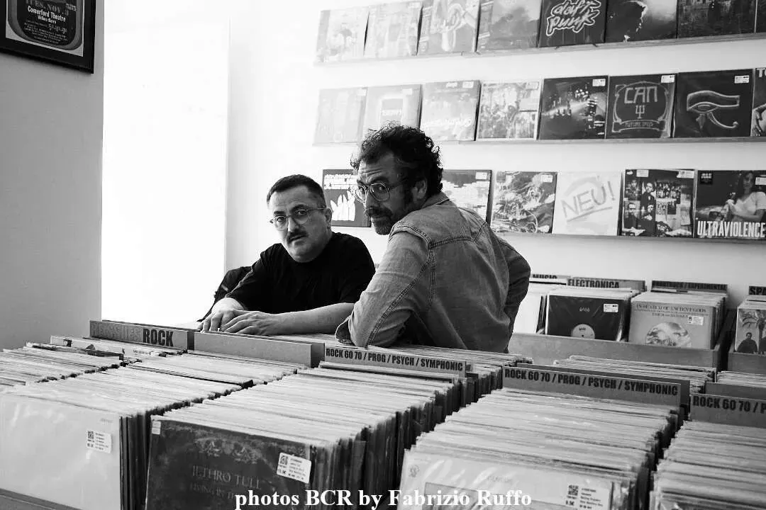 Dos hombres están sentados en una mesa en una tienda de discos mirando discos.