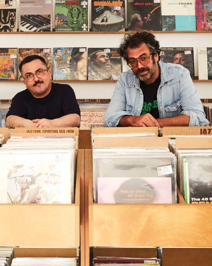 Dos hombres están sentados frente a un estante lleno de discos.