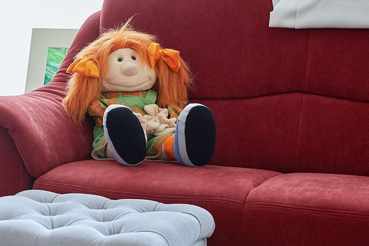 Eine orangehaarige Puppe sitzt auf einem roten Sofa neben einem hellblauen Hocker.