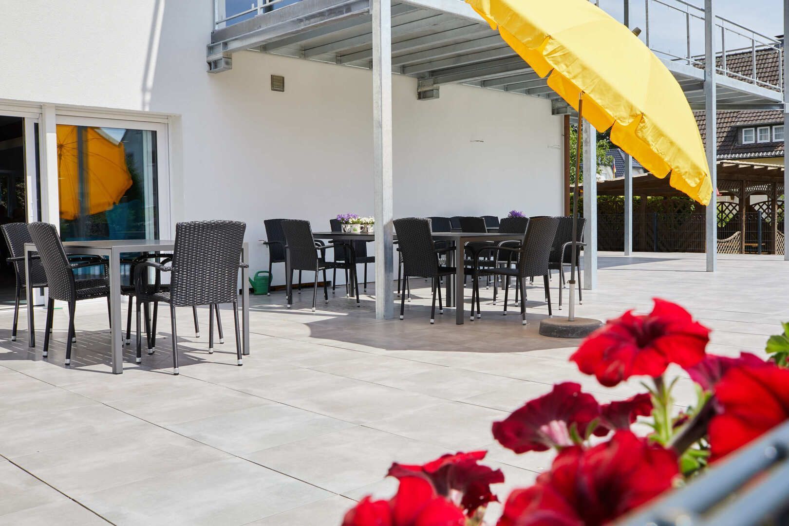 Terrasse eines Cafés im Freien mit schwarzen Tischen und Stühlen, weißem Gebäude, gelbem Sonnenschirm und roten Blumen im Vordergrund